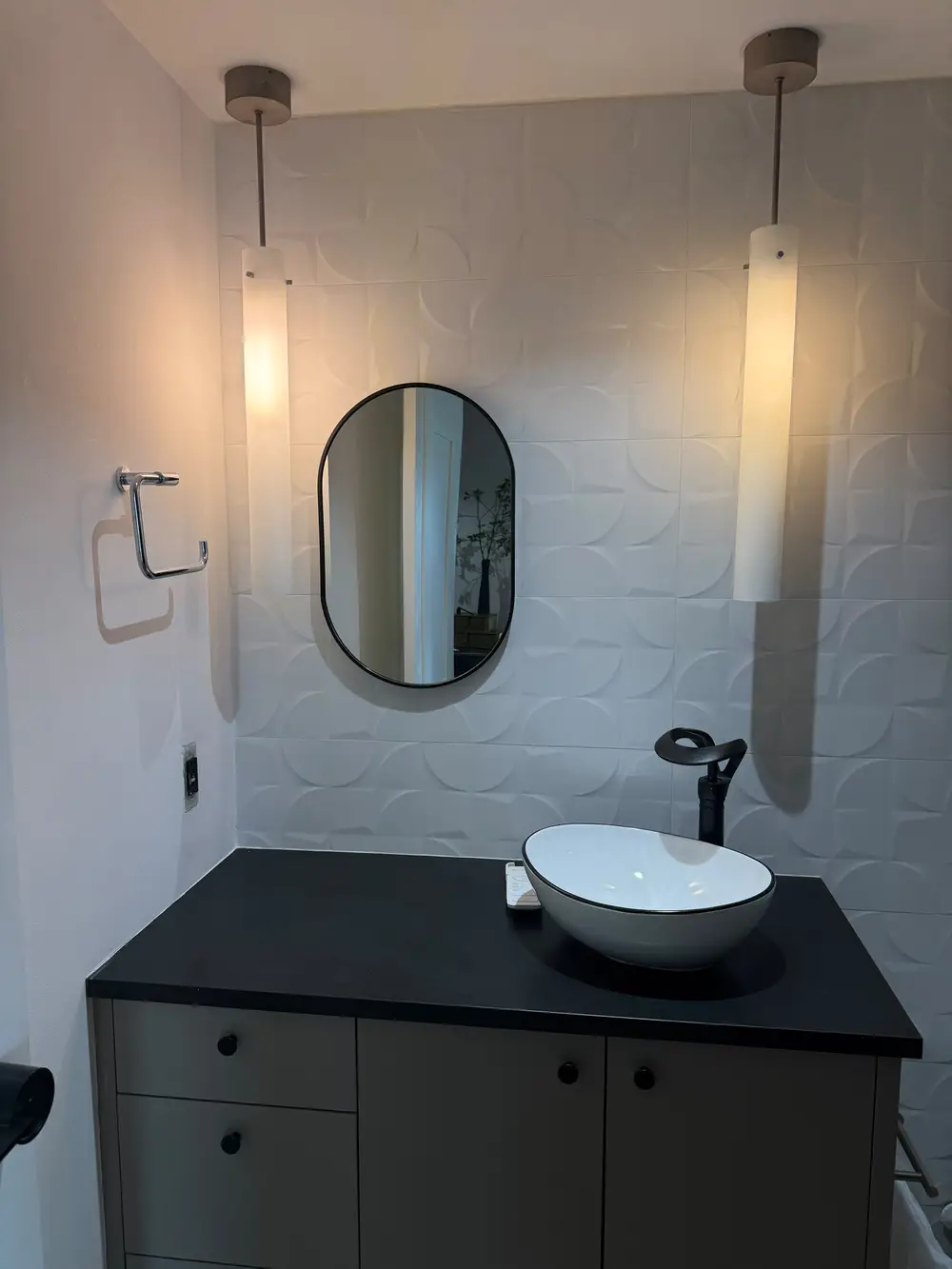 Modern powder room remodel by Evolve Design at the SW River Parkway project in Portland, featuring a textured white accent wall, an oval mirror, a white vessel sink on a black countertop, and contemporary pendant lighting