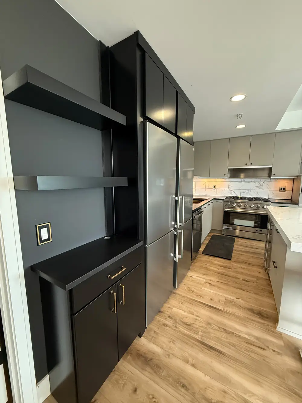 View of modern kitchen appliance integration by Evolve Design at SW River Parkway, Portland. Features a dark grey wall with black cabinetry and floating shelves alongside stainless steel appliances, with the warm-toned main kitchen area and marble backsplash visible in the background