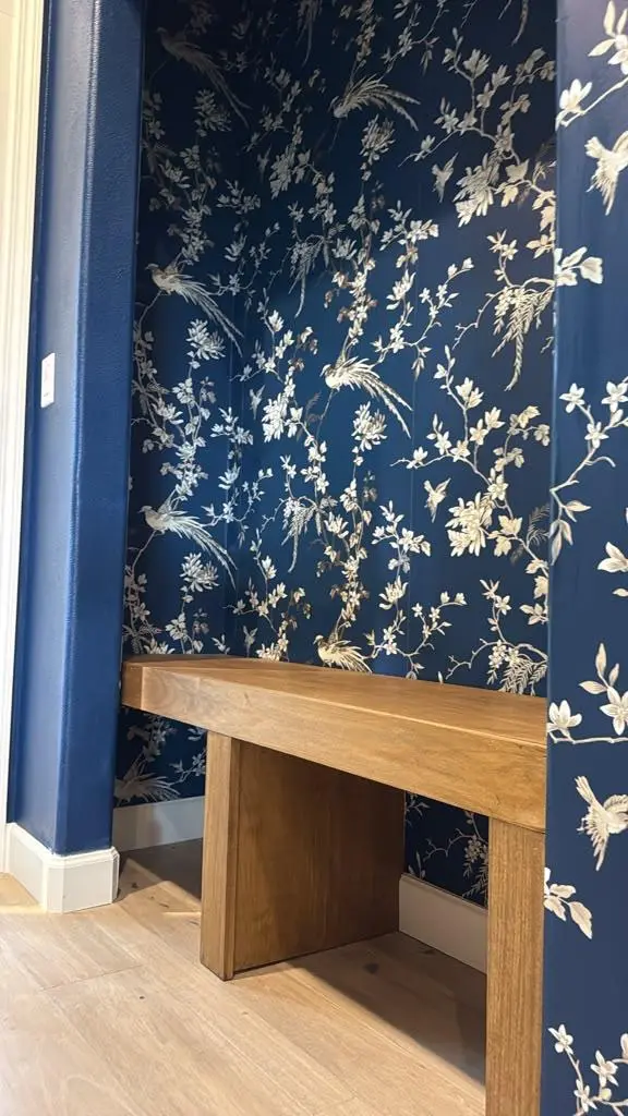 Angled view of a custom built-in entryway alcove designed by Evolve Design at the SW 14th St project in Gresham, Oregon. Features a solid natural wood bench set within a dark blue floral and bird motif wallpapered alcove, finished with crisp white baseboards and light hardwood flooring
