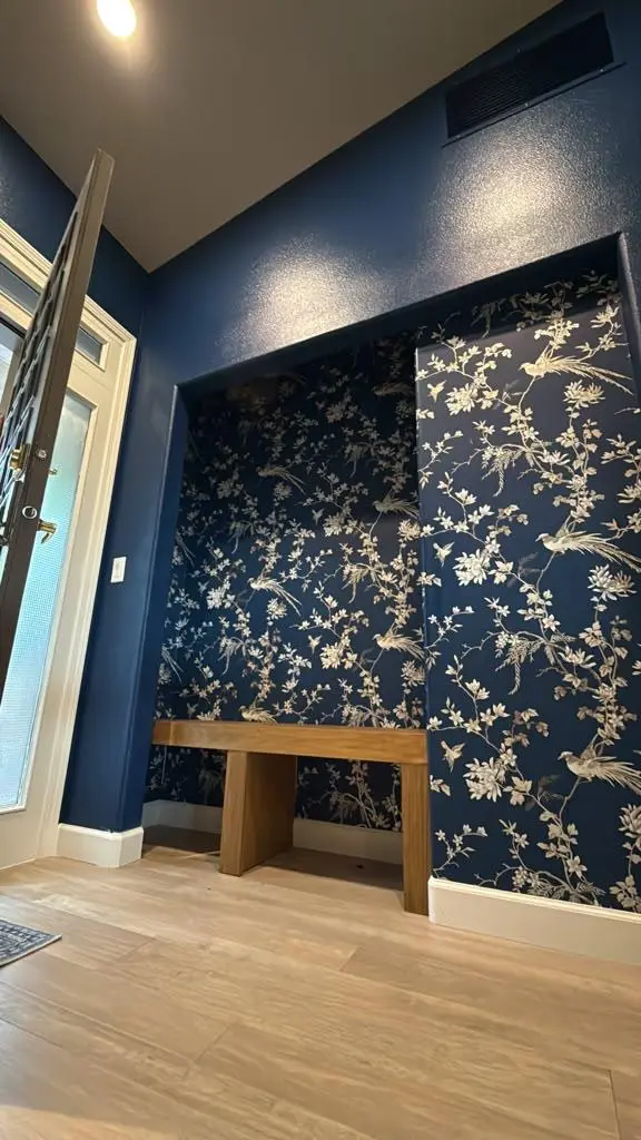 Wide low-angle view of a custom entryway foyer remodel by Evolve Design at SW 14th St in Gresham, Oregon. It showcases a modern open front door leading into a hallway with light hardwood floors, a deep blue painted wall, and a recessed alcove with a natural wood bench against an elegant navy and gold floral wallpaper