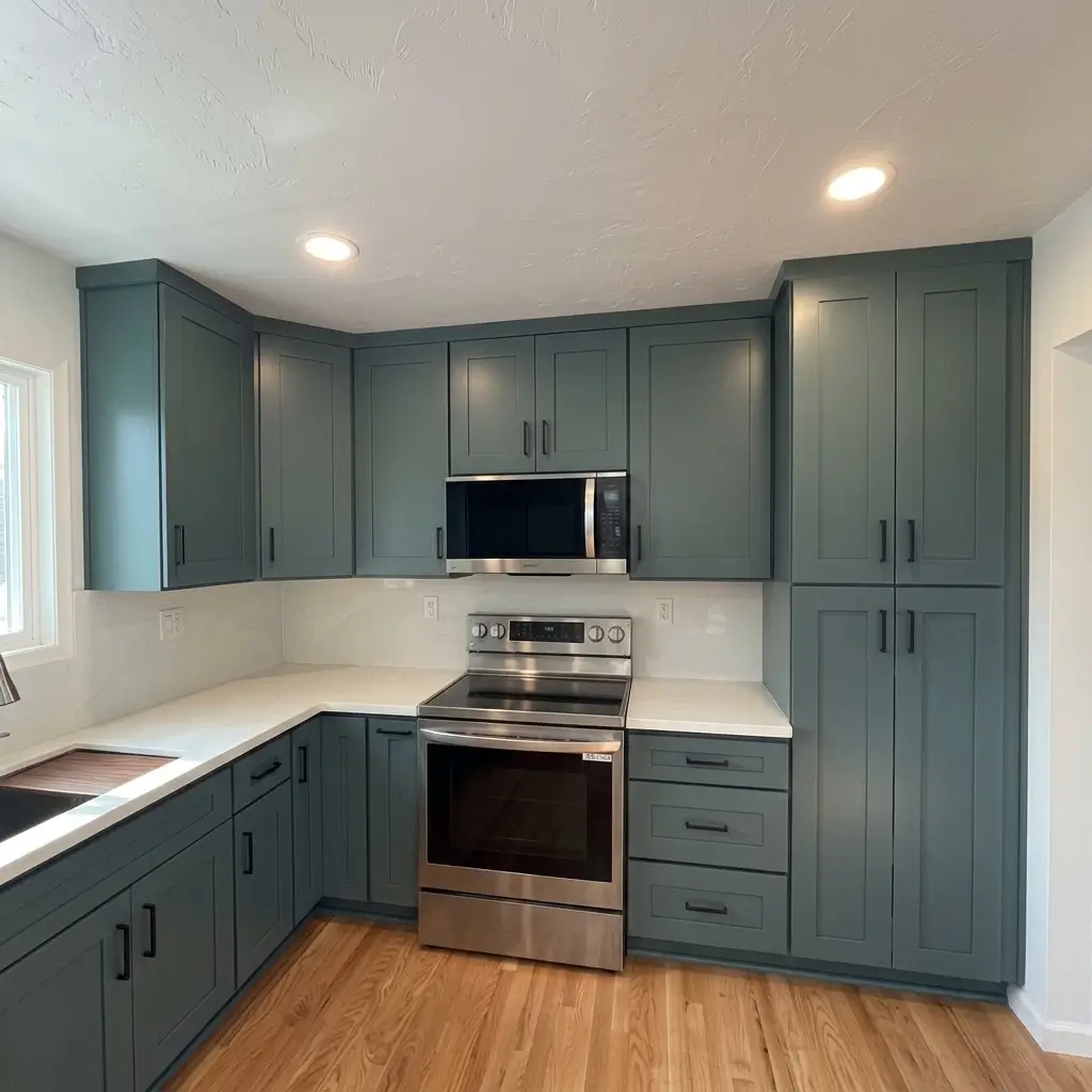 Completed kitchen remodeling transformation in Damascus, Oregon, featuring dark teal shaker cabinetry, white solid-surface countertops, stainless steel appliances, and natural hardwood floors
