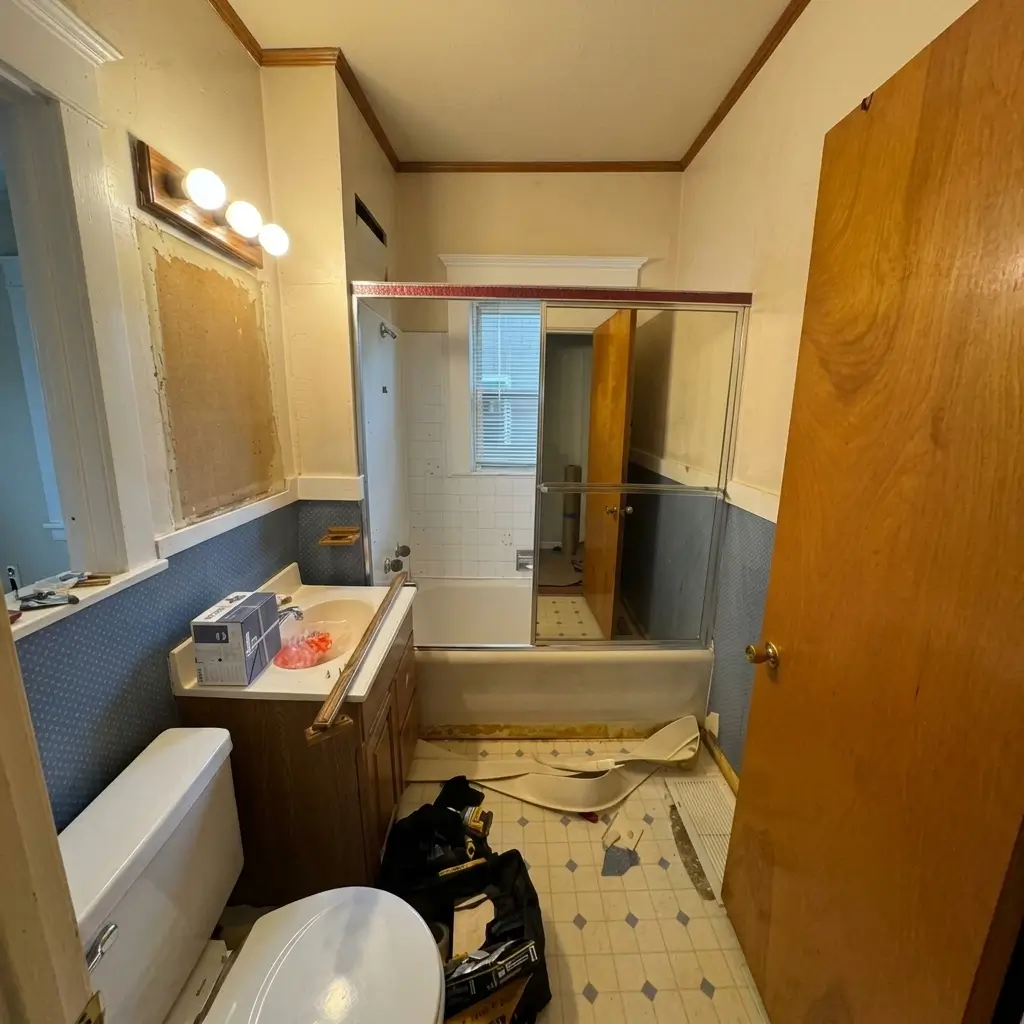 Wide-angle view of a Portland bathroom during early renovation prep, showing contractor tools, partially removed fixtures, old blue wallpaper, and bathtub glass enclosure
