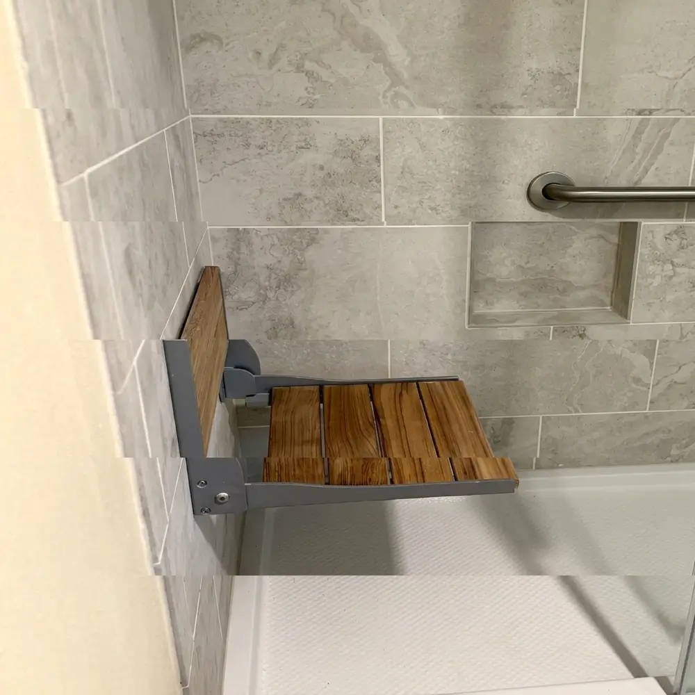 Wall-mounted fold-down teak shower bench inside a modern walk-in shower with gray marbled tiles, recessed niche, and stainless steel grab bar in a Woodland, Washington home