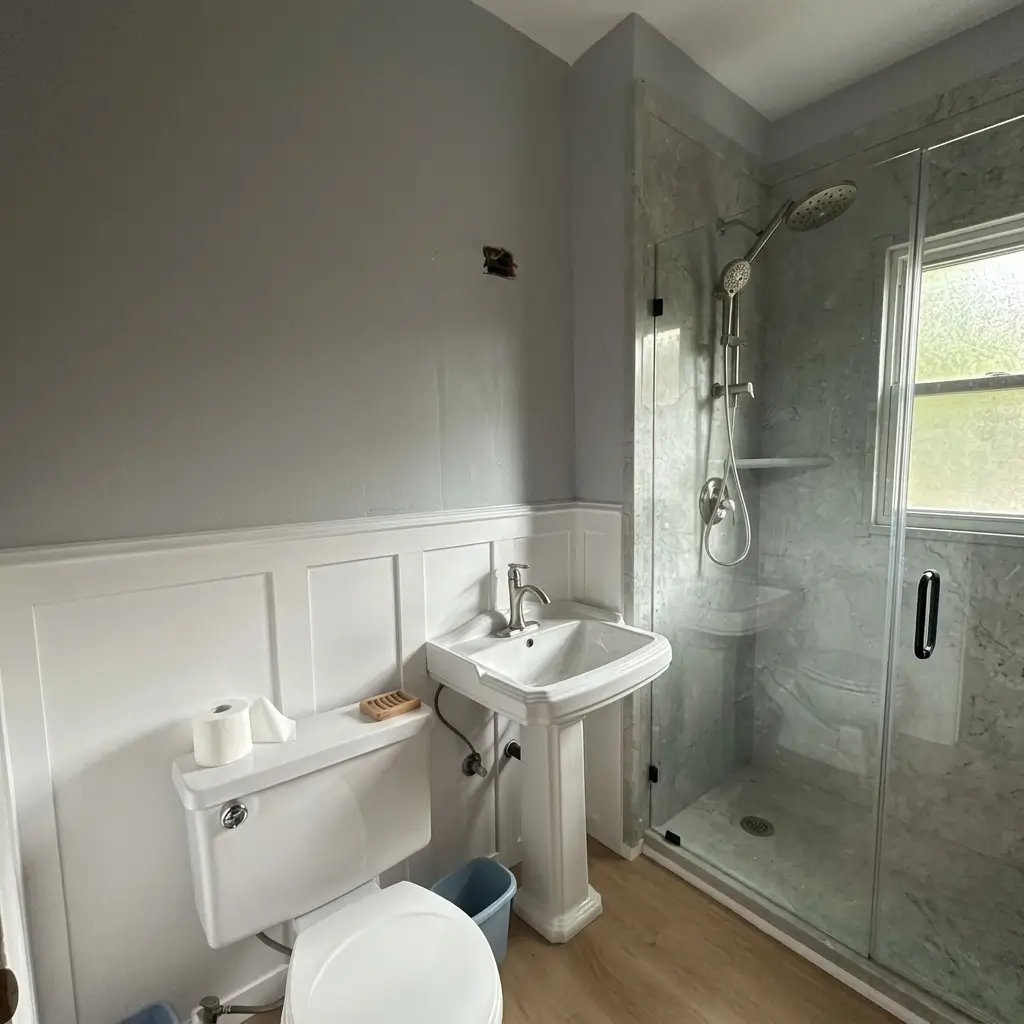 Close-up view of a classic white pedestal sink, toilet, white wainscoting paneling, and sliding glass shower door in a remodeled Portland bathroom