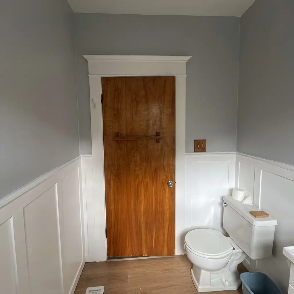 Natural wood interior door framed by white trim, flanked by white wainscoting wall paneling and grey walls in a remodeled Portland bathroom
